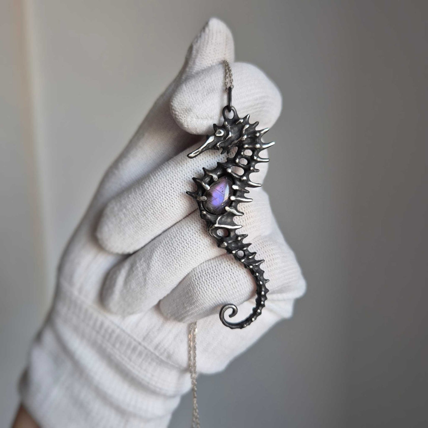 Close-up of silver seahorse necklace in a gloved hand, highlighting its curved shape and shimmering purple labradorite