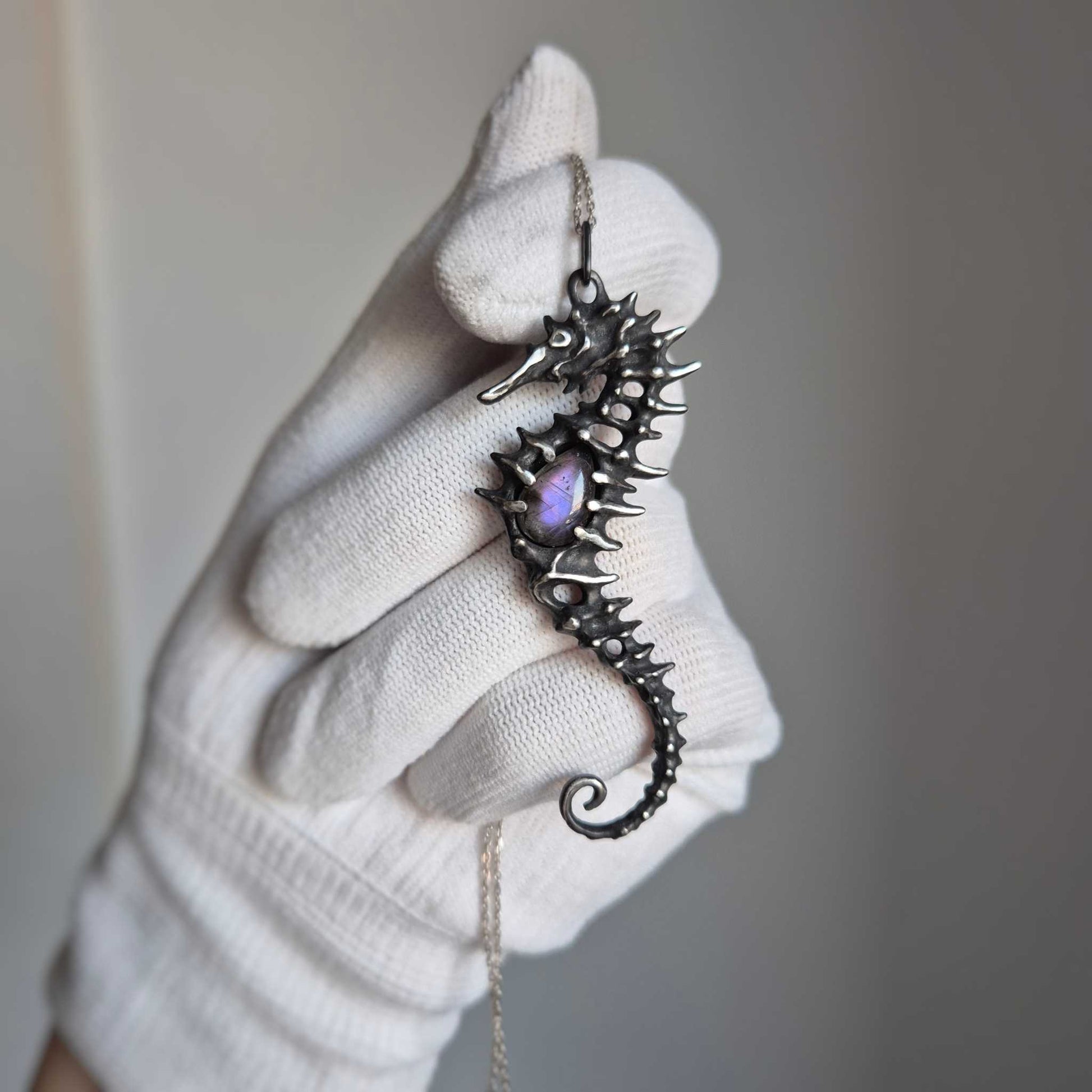Close-up of silver seahorse necklace in a gloved hand, highlighting its curved shape and shimmering purple labradorite