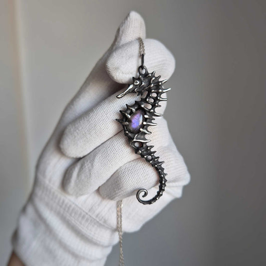 Close-up of silver seahorse necklace in a gloved hand, highlighting its curved shape and shimmering purple labradorite