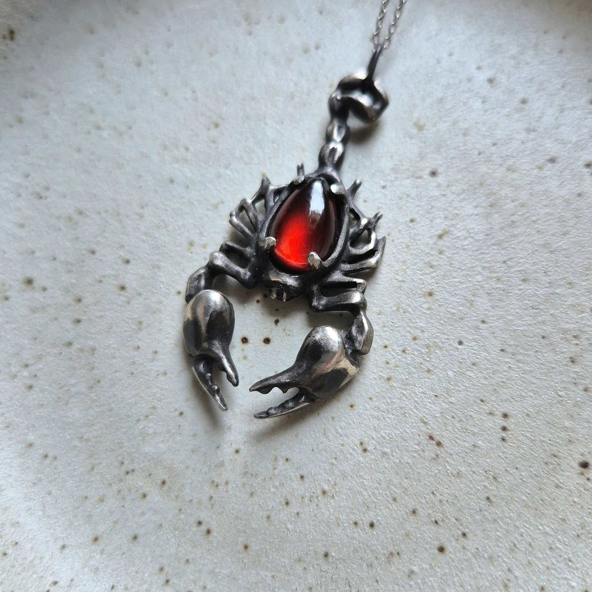 Detailed shot of the Scorpion necklace featuring a glowing red cabochon gem set in oxidized silver, shown on a minimalist ceramic surface.