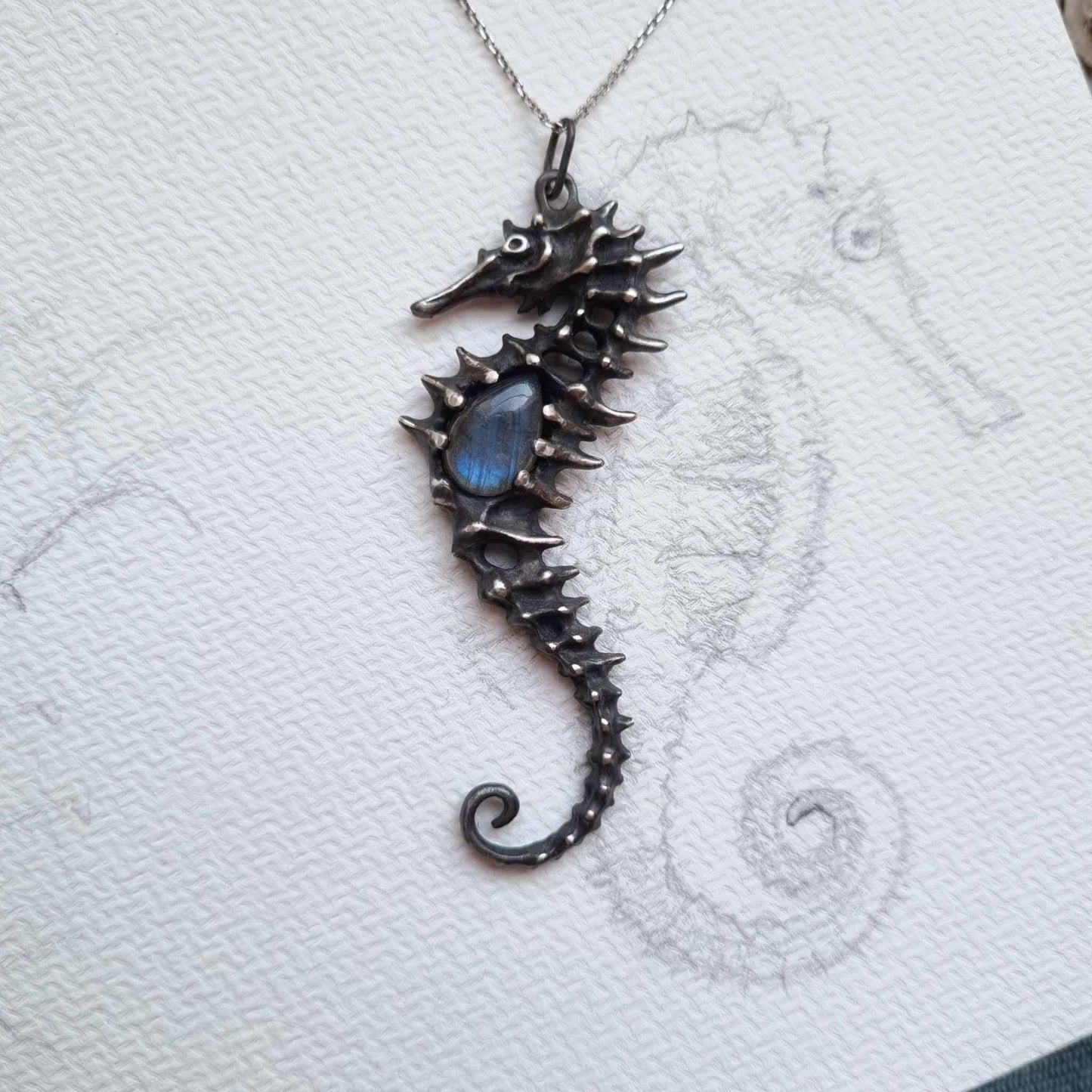 Silver Seahorse necklace with sculpted texture and blue labradorite on textured paper background with seahorse sketch.