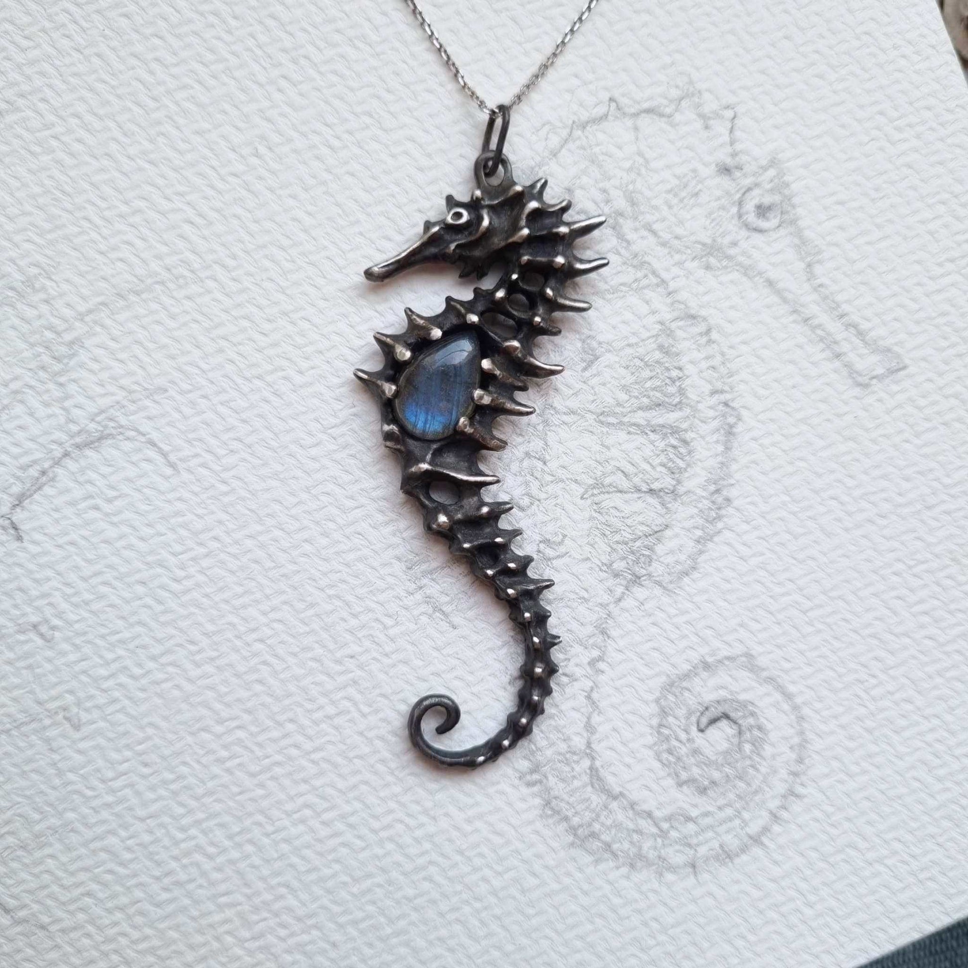 Silver Seahorse necklace with sculpted texture and blue labradorite on textured paper background with seahorse sketch.