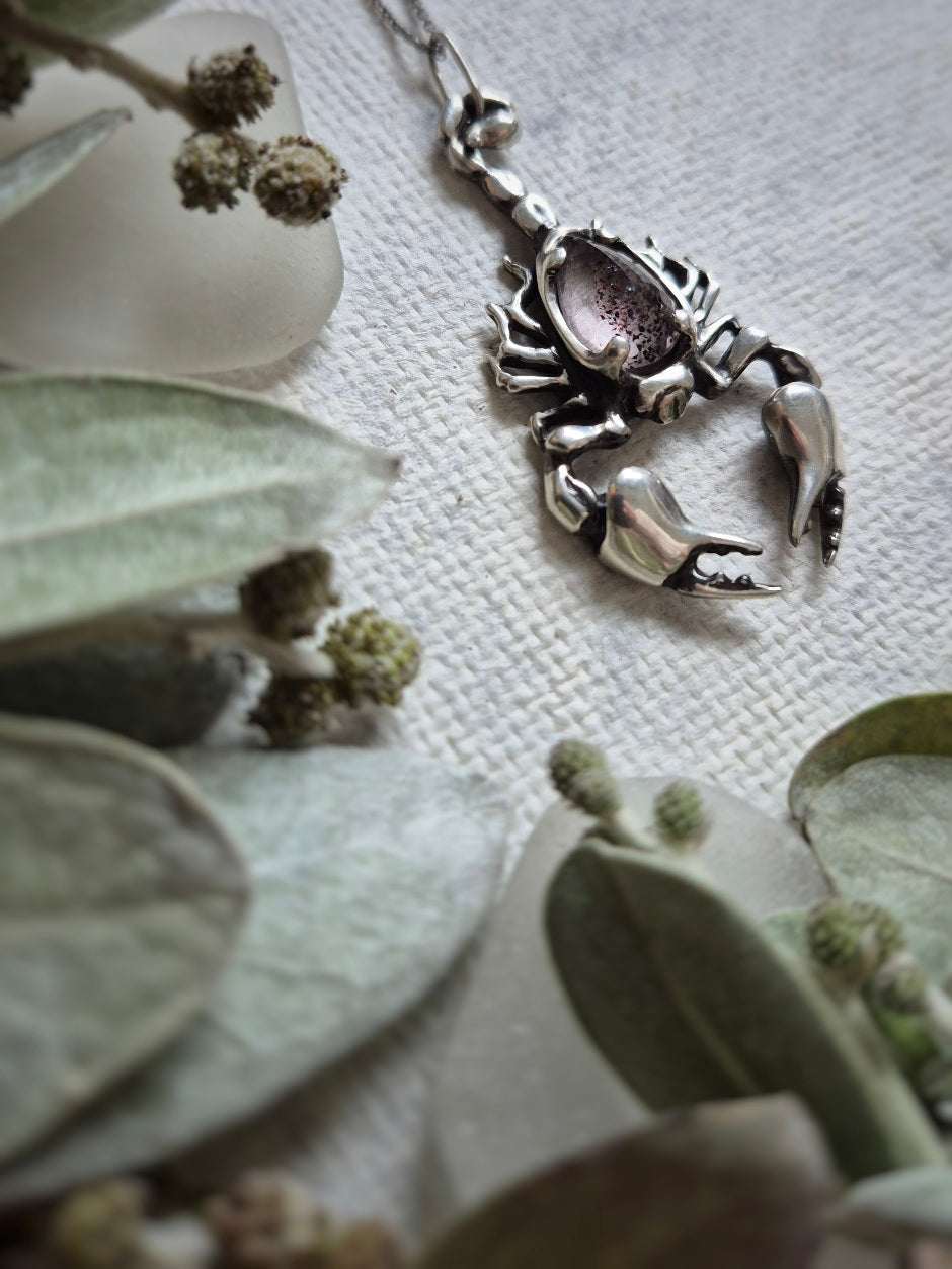Angled shot of the silver Scorpion necklace nestled in foliage, emphasizing the gem's unique inclusions.
