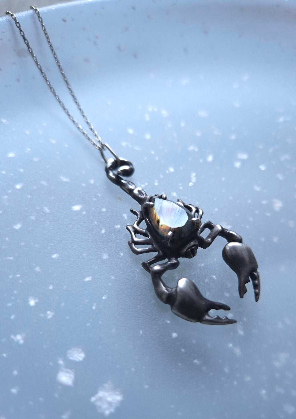 Silver Scorpion necklace with a faceted labradorite, photographed against a blue textured background.