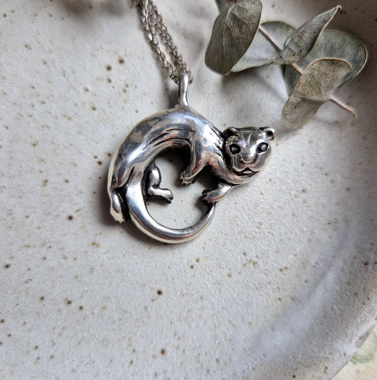Close-up of the silver Otter necklace on a neutral ceramic background, highlighting the curved, sculptural shape of the pendant.