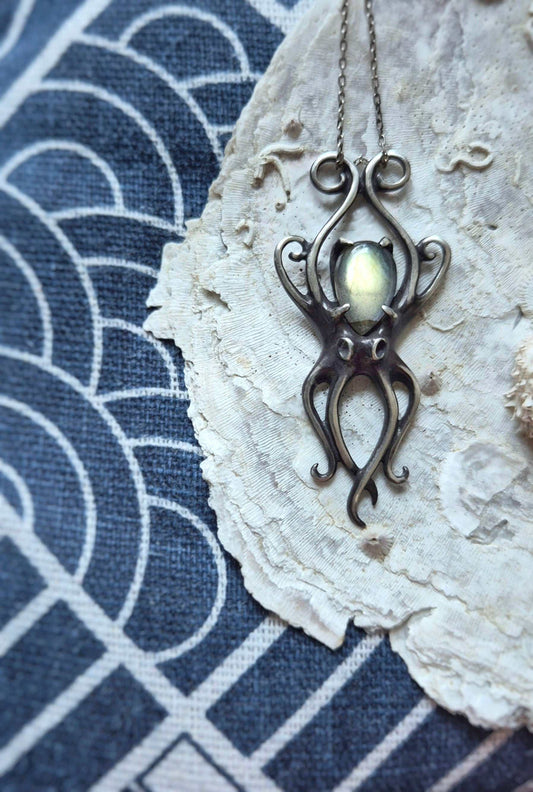 Handmade silver Octopus necklace with glowing labradorite gemstone, photographed on textured shell and indigo fabric background with ethnic pattern