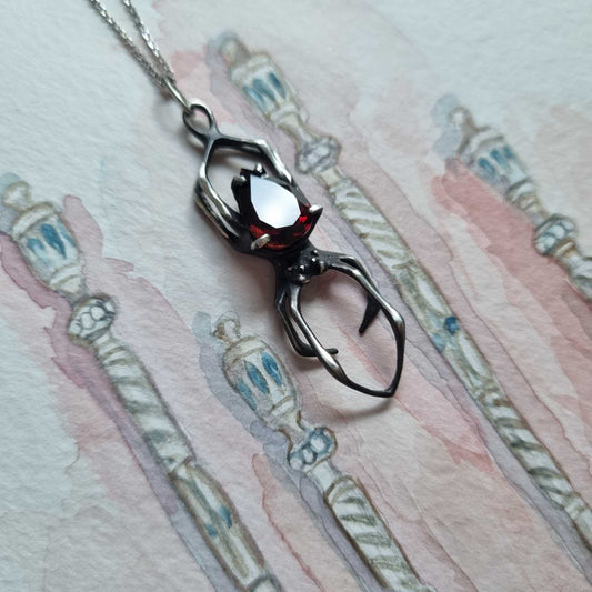 Oxidized silver spider necklace with faceted garnet resting on a watercolor jewelry sketch