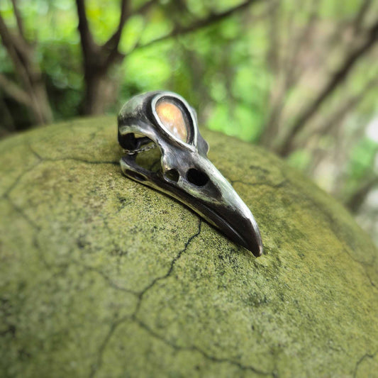 Raven skull pendant featuring golden labradorite “third eye,” photographed on green mossy rock surface.