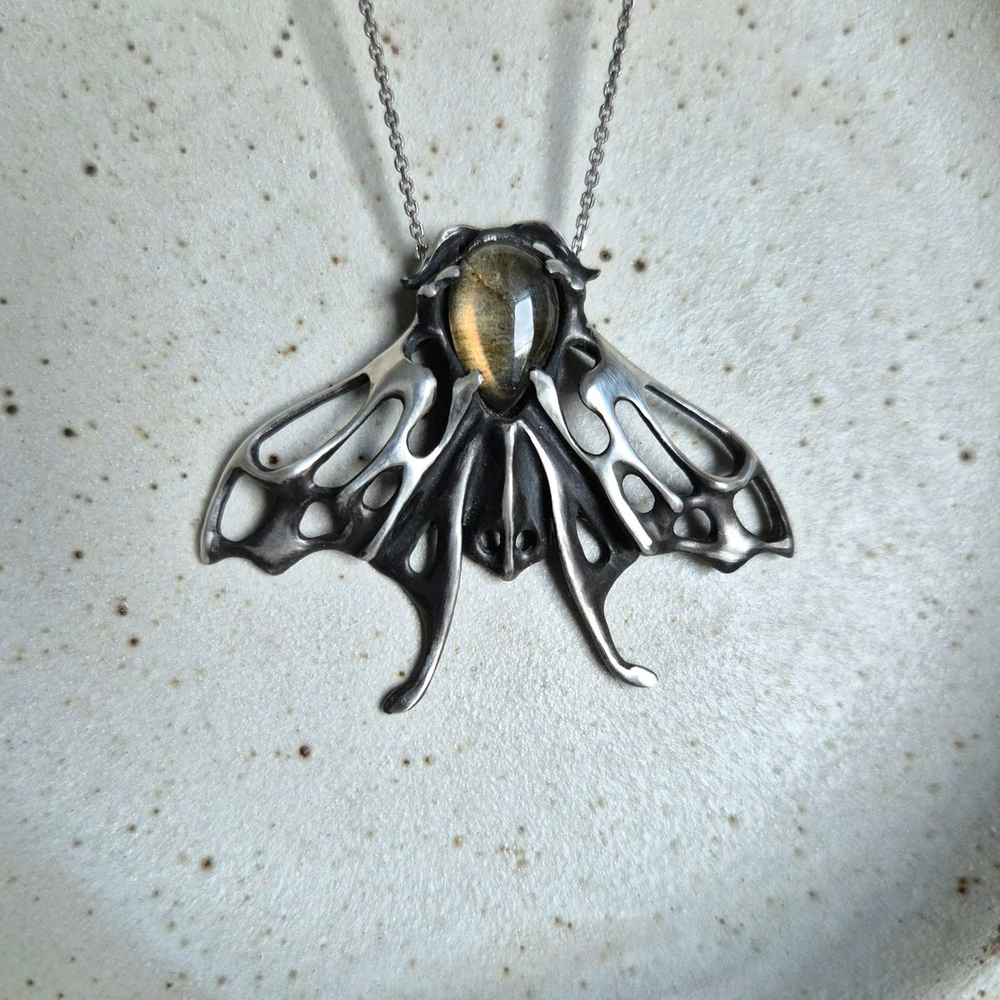 Sculptural silver Moth pendant with glowing yellow labradorite, against neutral ceramic background
