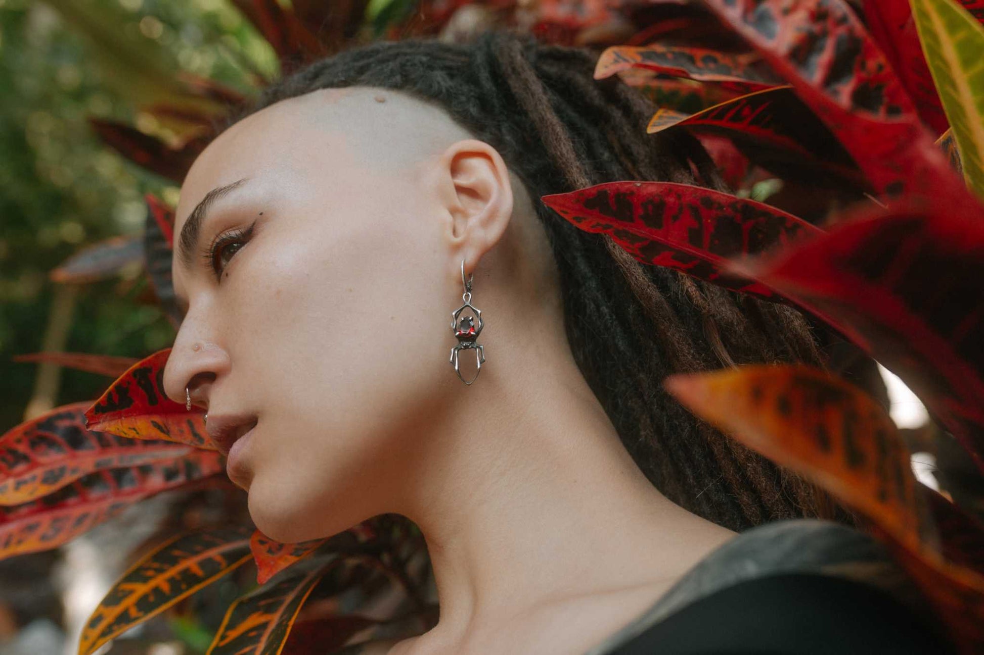 Handmade silver spider earrings with natural garnets, worn by a model with dreadlocks in a shamanic-inspired look, set against a background of natural leaves