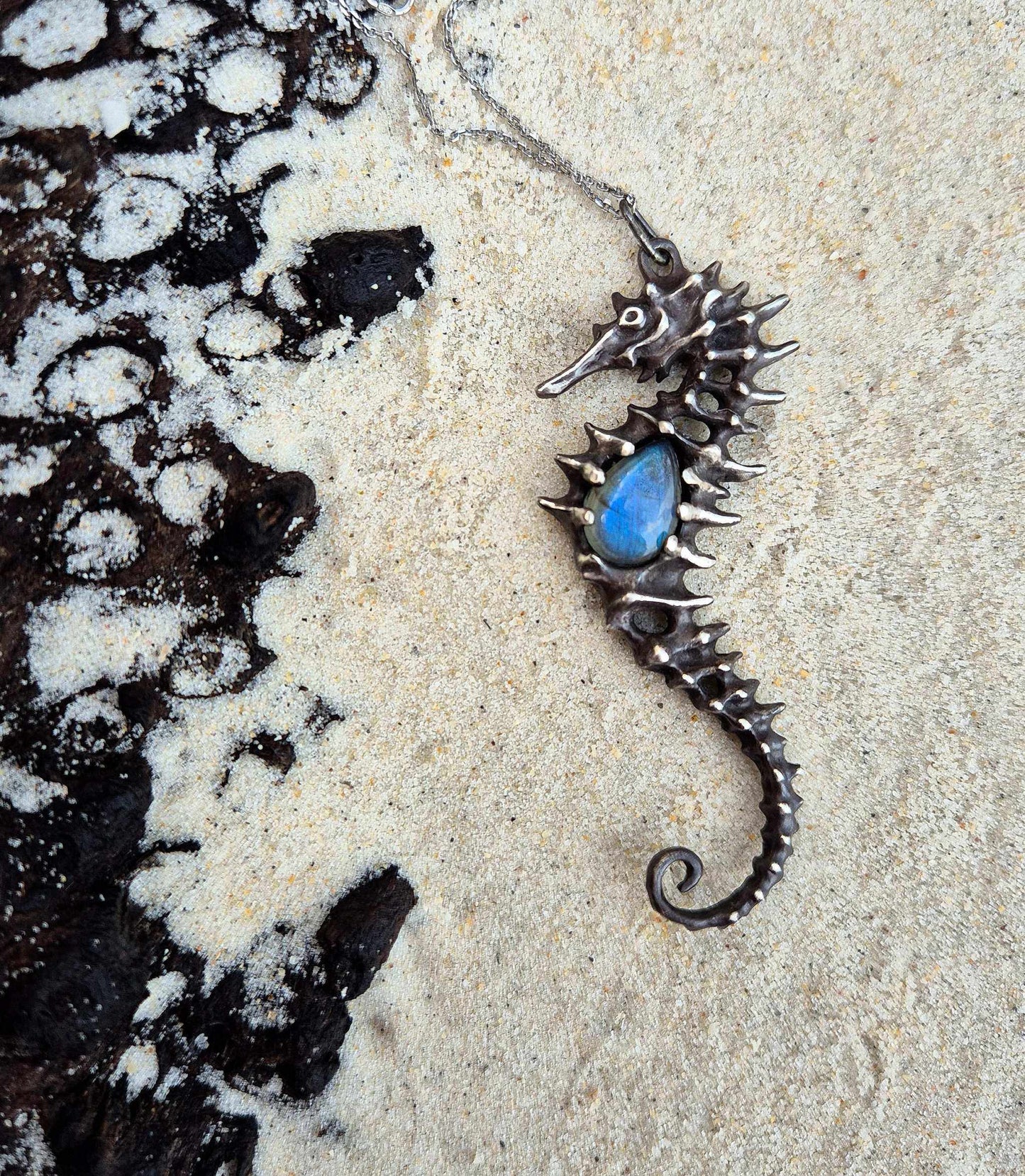 Oxidized silver Seahorse necklace with labradorite center, resting on sandy beach near black volcanic rock.