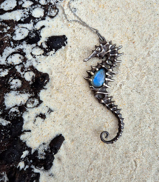 Oxidized silver Seahorse necklace with labradorite center, resting on sandy beach near black volcanic rock.