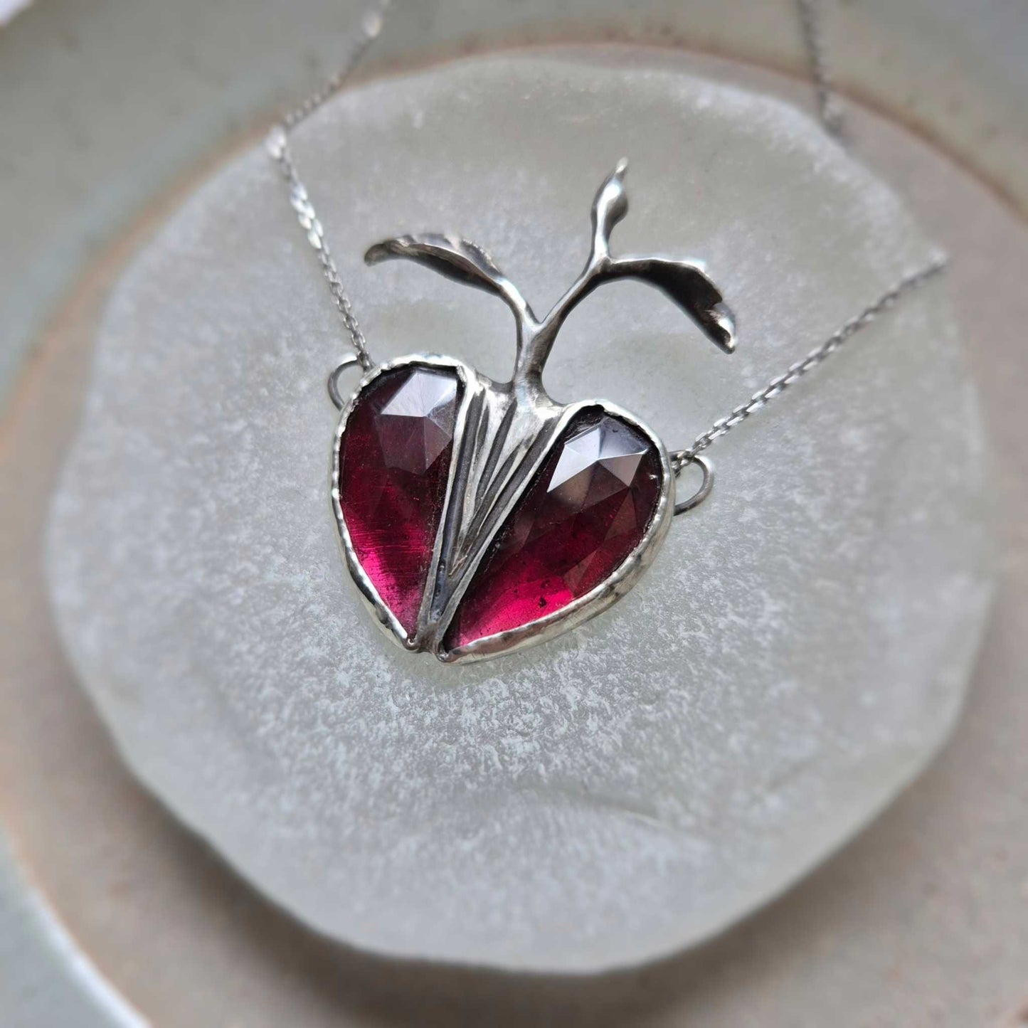 Silver Seed Heart necklace photographed from above, showing the symmetry of the garnet stones on a round white display.