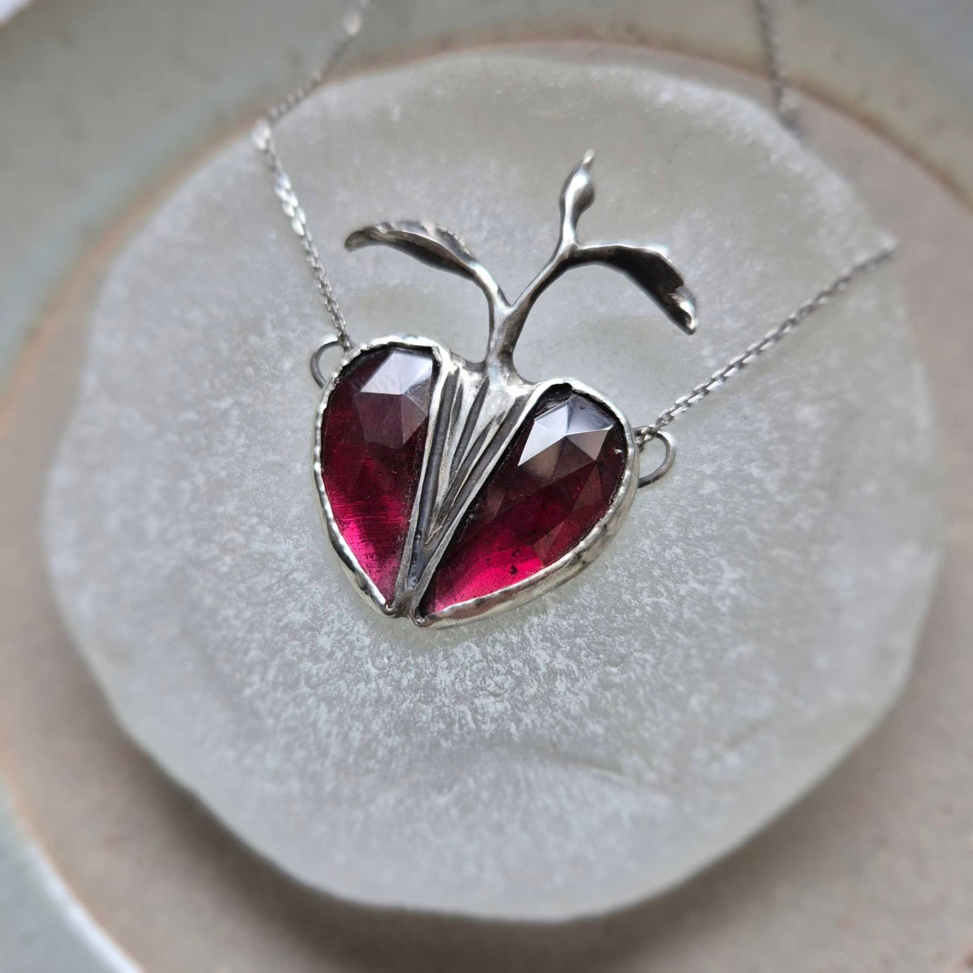 Silver Seed Heart necklace photographed from above, showing the symmetry of the garnet stones on a round white display.