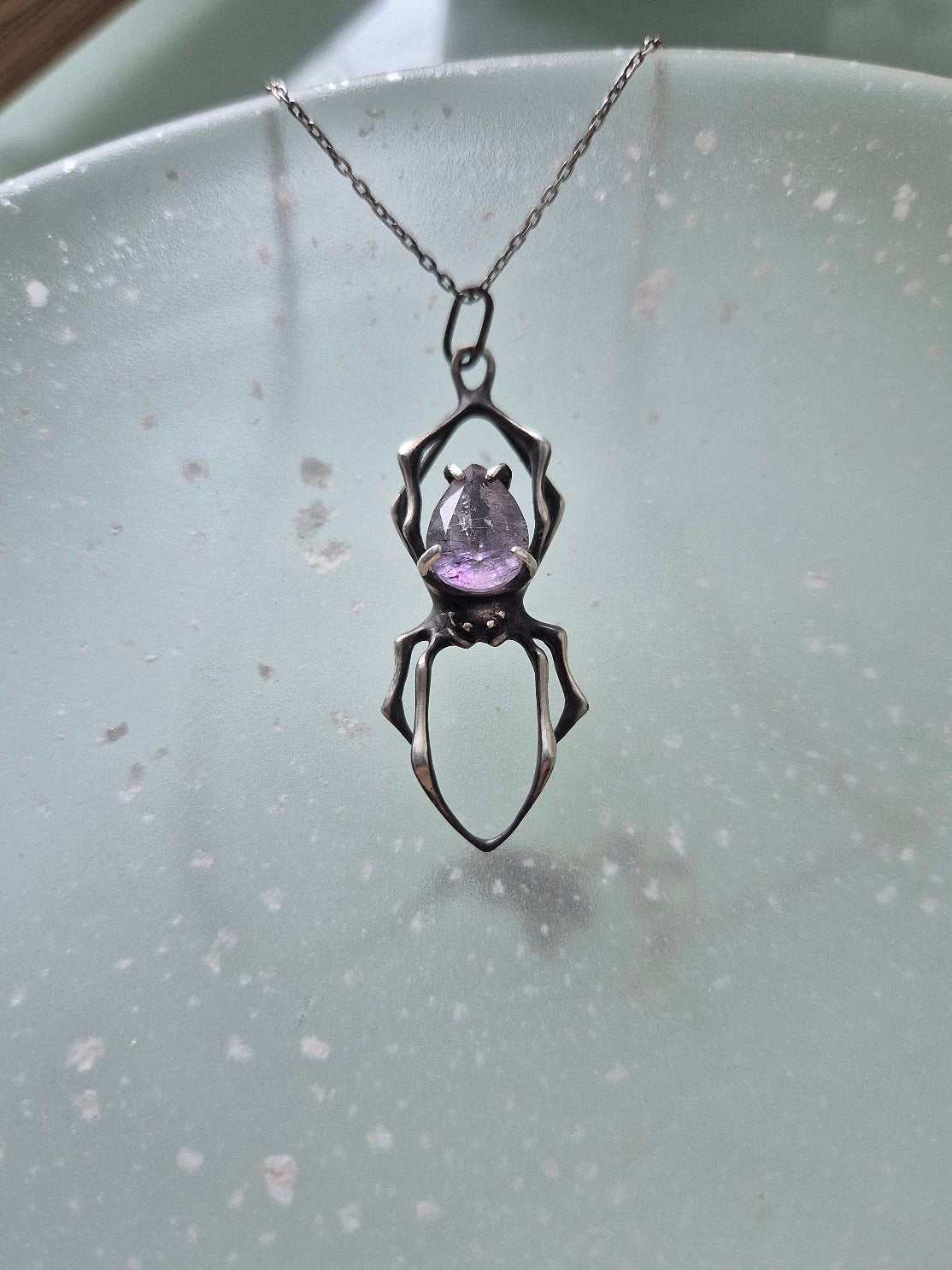 Close-up of a silver spider pendant with a raw-cut purple Super Seven on a translucent surface with scattered reflections