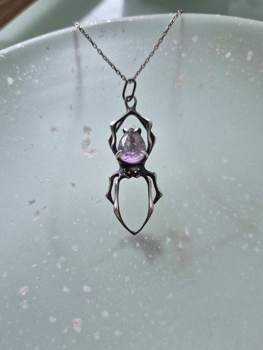 Close-up of a silver spider pendant with a raw-cut purple Super Seven on a translucent surface with scattered reflections