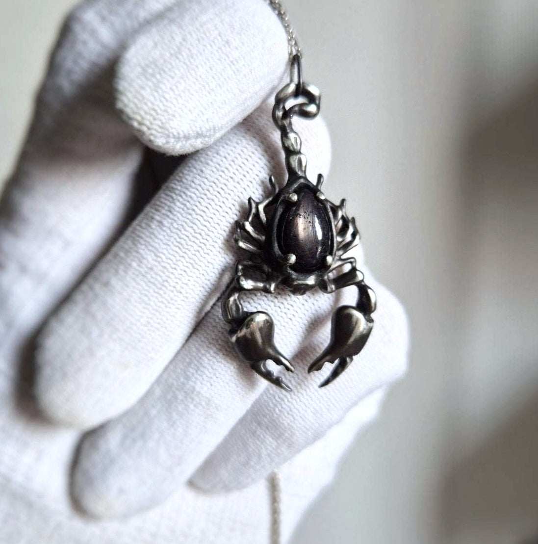 gloved hand holding silver Scorpion necklace to highlight black gemstone and sculptural design