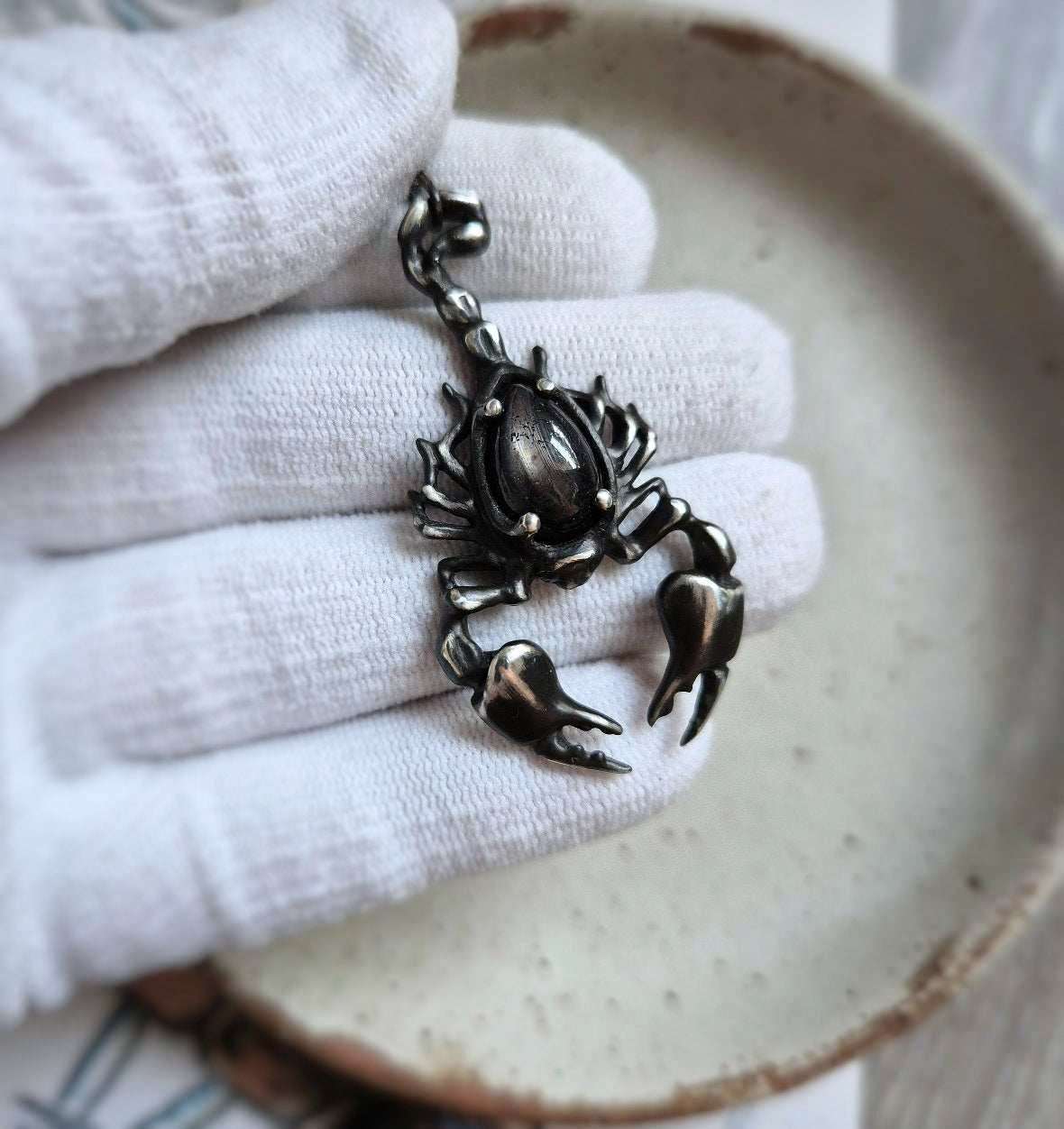 silver Scorpion pendant held in a gloved hand, showcasing details of the oxidized silver and black gemstone