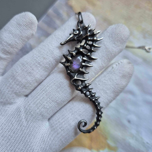 Hand holding a silver seahorse necklace with purple labradorite gemstone, shown against a pale background