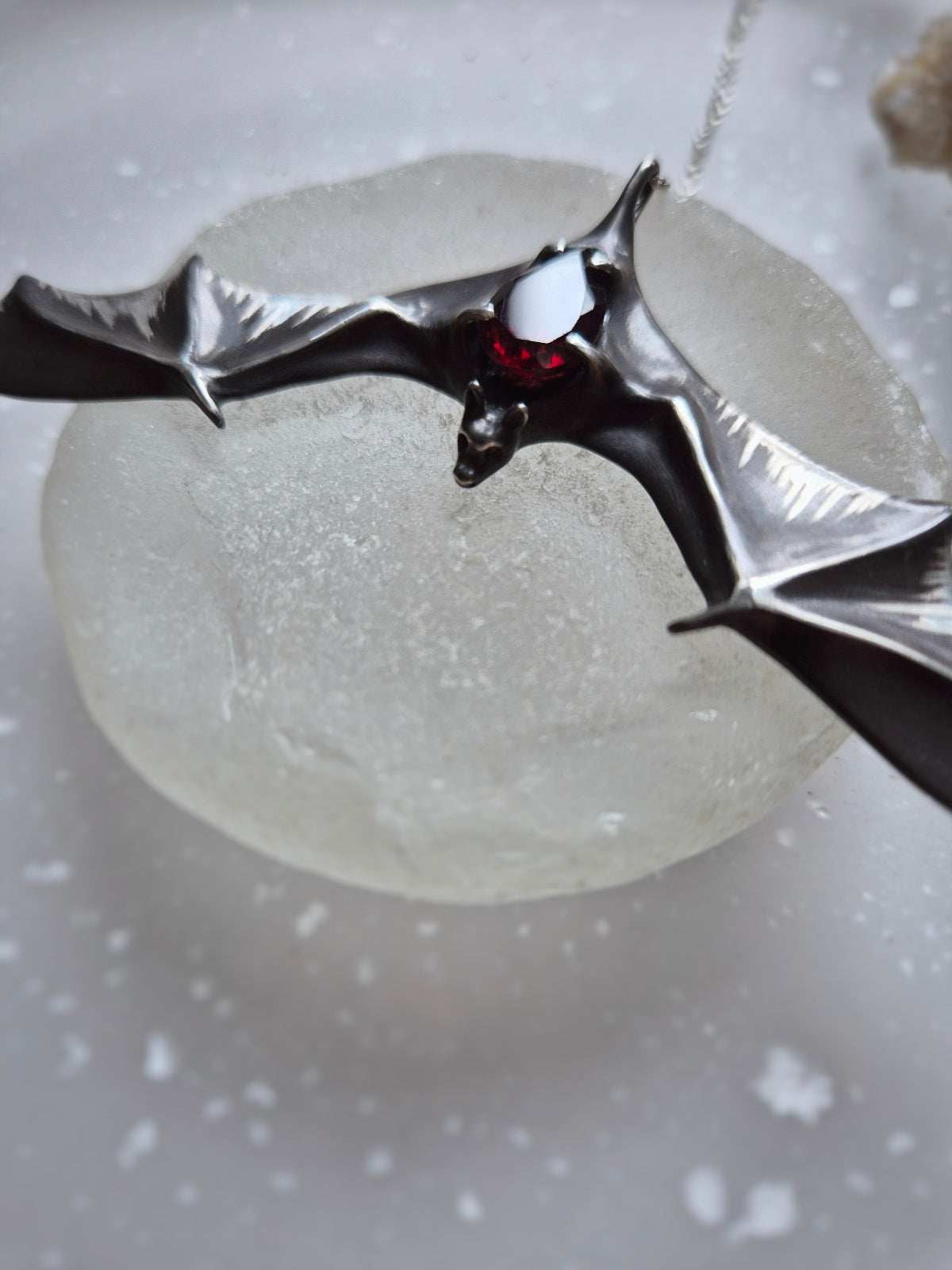 Close-up of Silver Bat necklace featuring a natural garnet gemstone, placed on a round frosted glass surface.