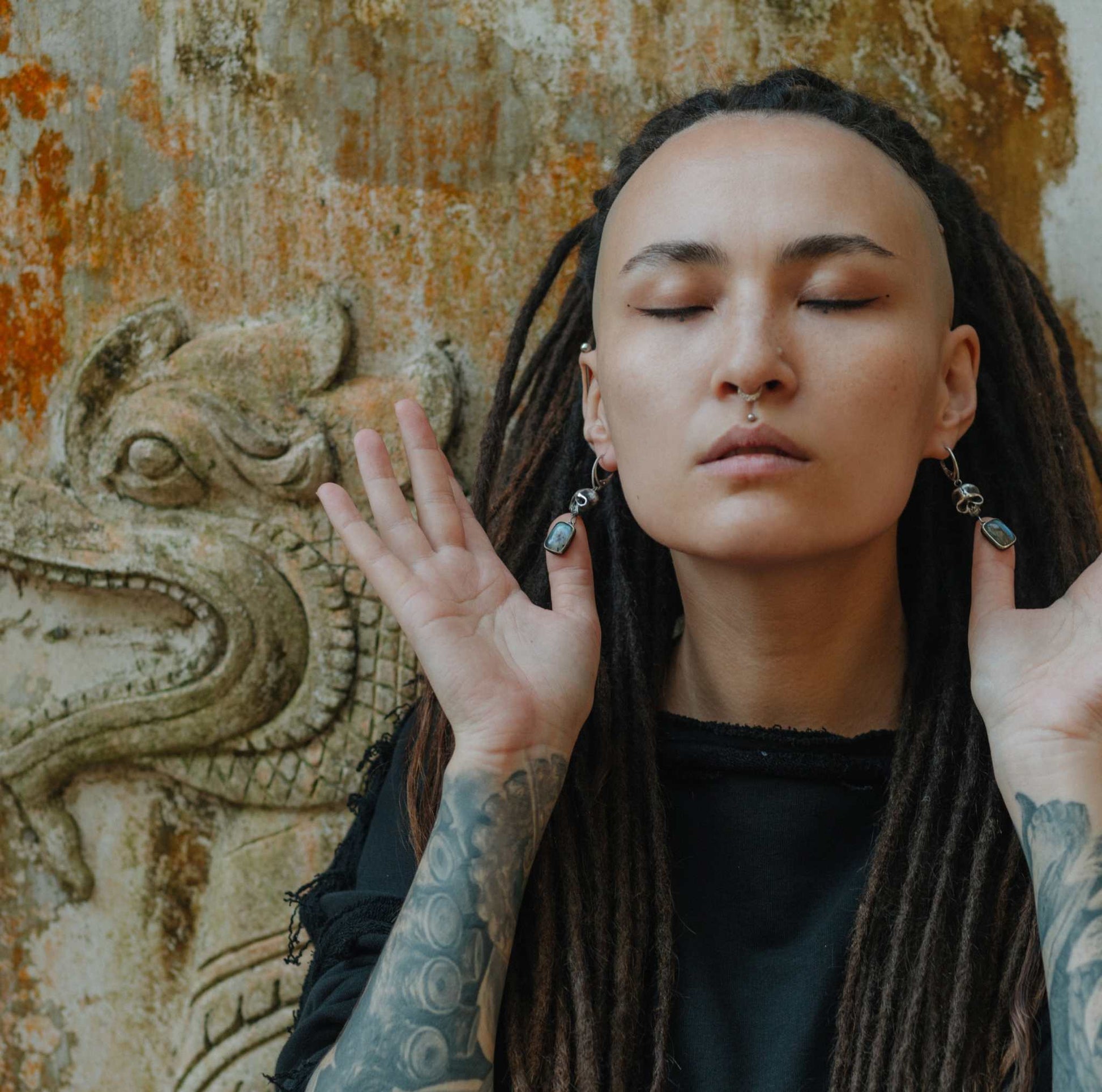 Oxidized silver skull earrings with labradorite gemstones, worn by a model with dreadlocks and closed eyes, against an ancient textured wall