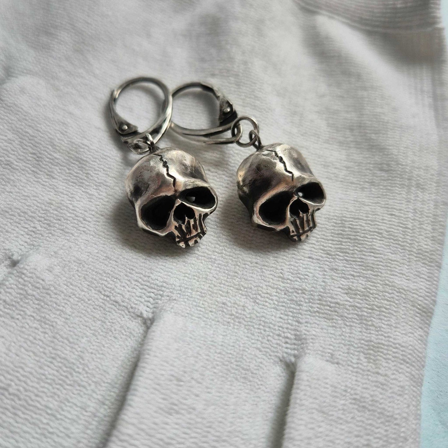Close-up of handcrafted silver Skull earrings resting on a white glove, showcasing their sculptural detail.