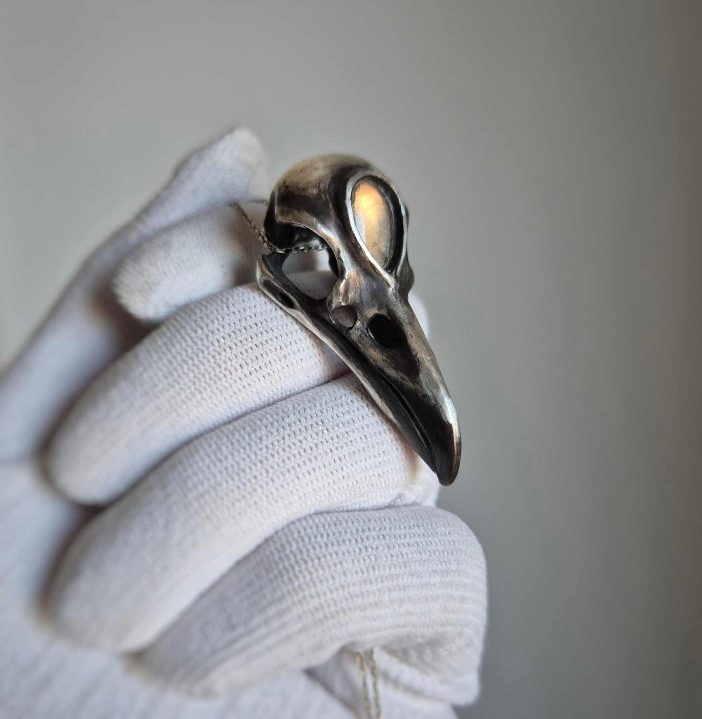 Silver raven skull necklace with golden labradorite, held in gloved hand for close-up detail.