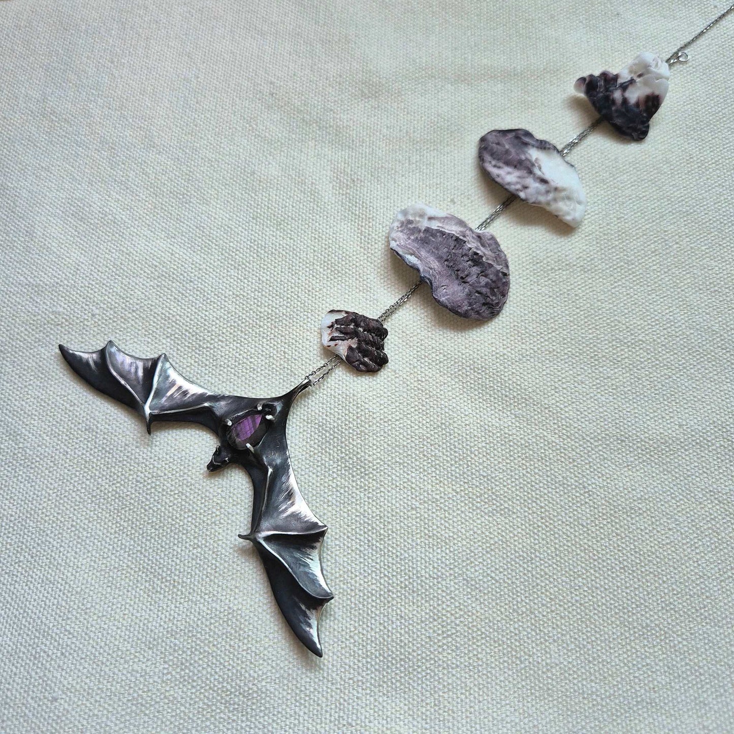 Sculptural silver Bat pendant with labradorite gem, laid out in full view on light fabric, and a chain decorated with seashells