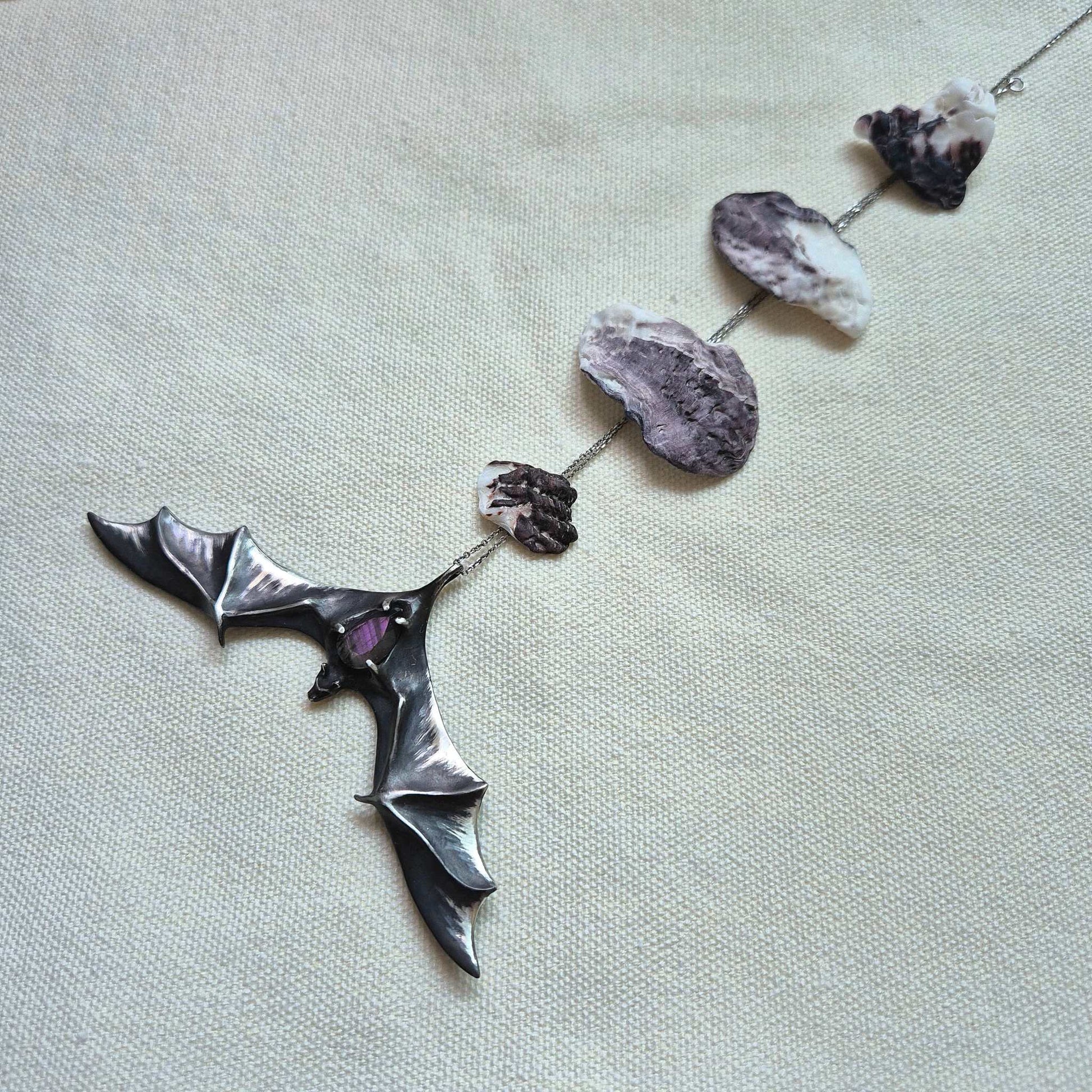 Sculptural silver Bat pendant with labradorite gem, laid out in full view on light fabric, and a chain decorated with seashells