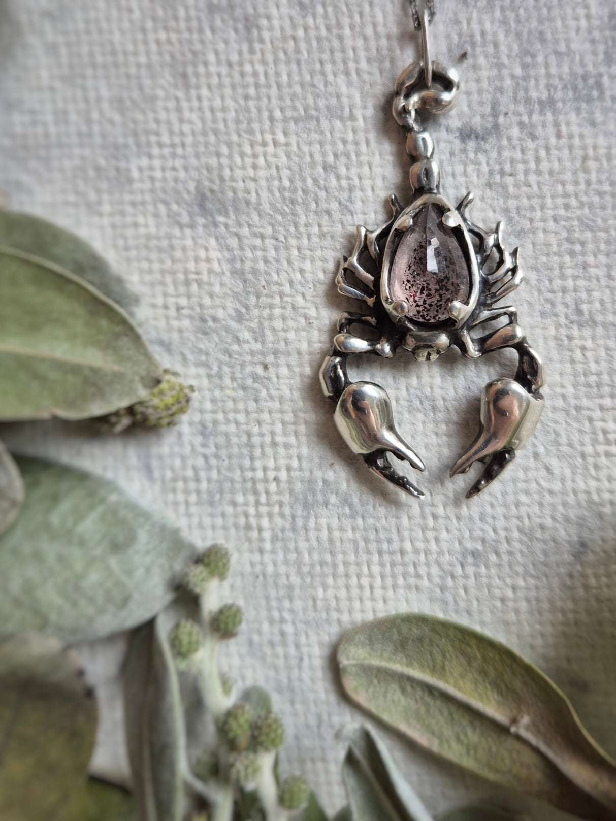 Hanging view of the Scorpion pendant surrounded by plants, showcasing its weight and intricate design.