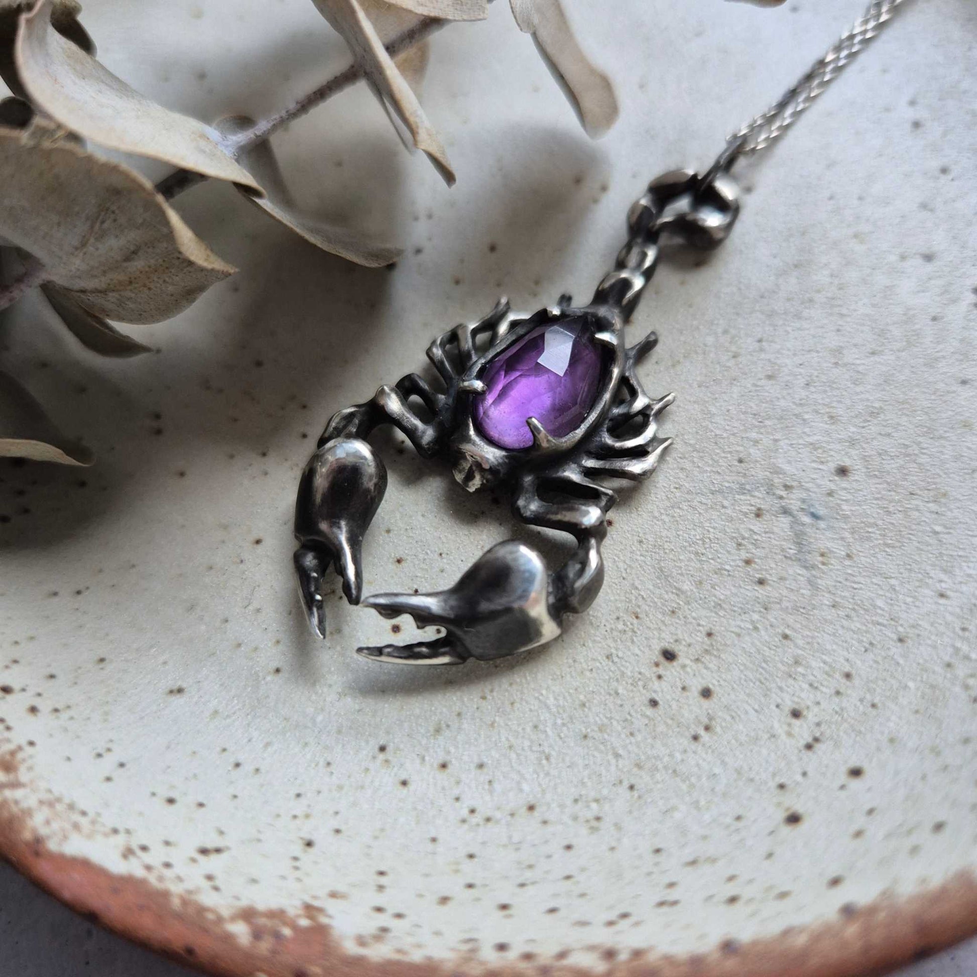 Top view of a silver Scorpion necklace with an amethyst, on ceramic background with botanical decor around