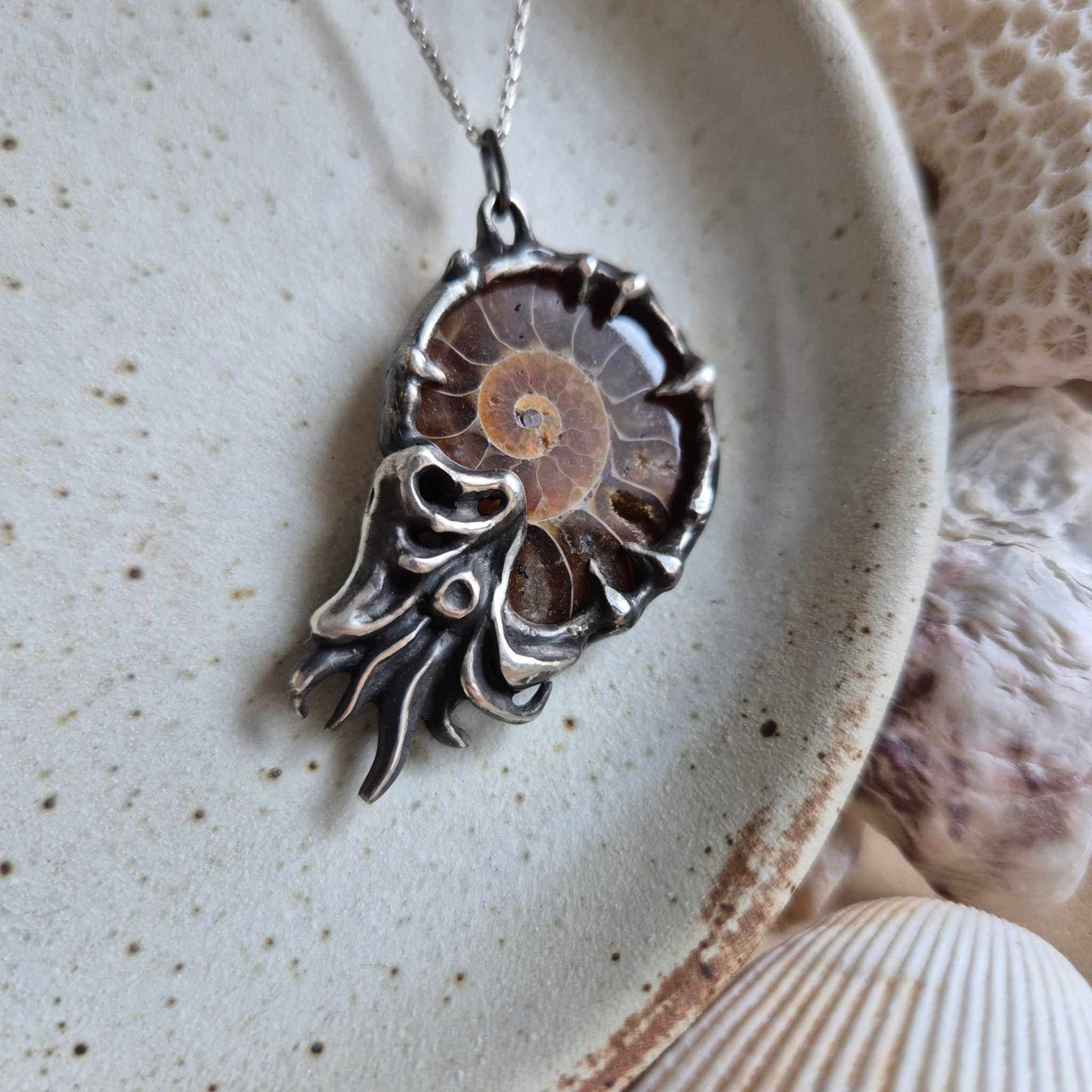 Handmade ышдмук Nautilus necklace in oxidized silver with an ammonite, resting on a textured ceramic surface among ocean-themed decor.