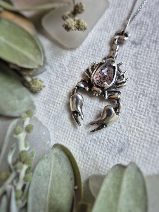 Close-up of silver Scorpion necklace with Super Seven gemstone, styled on soft green foliage.