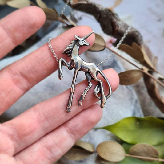 Silver Unicorn necklace shining under natural light, displayed on a hand for scale, with green leaves on the background