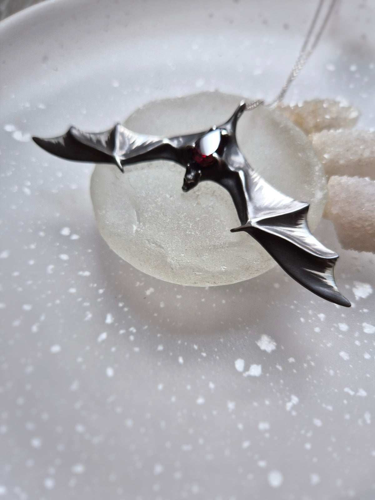 Silver Bat necklace lying on a translucent textured background, showcasing the garnet’s subtle glow.