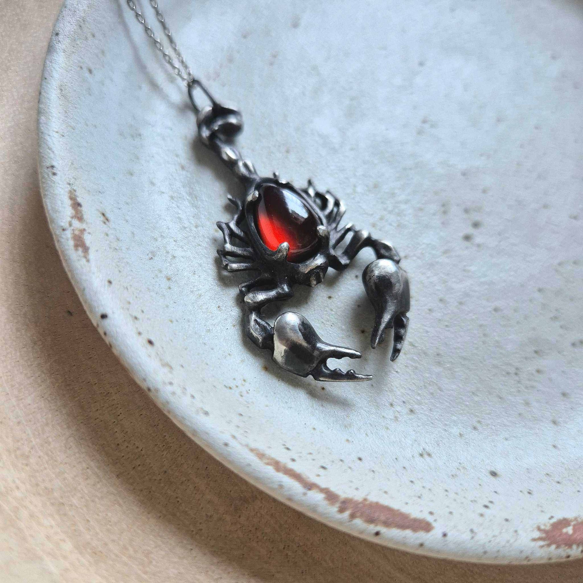 Side view of the red gemstone silver Scorpion pendant, highlighting its curved claws and organic silver structure against a rustic plate.