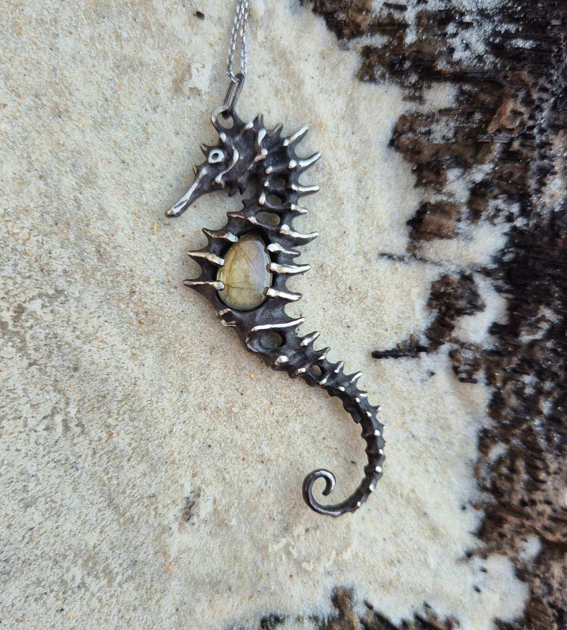 Oxidized silver Seahorse necklace with a gemstone center, resting on sandy beach near black volcanic rock.