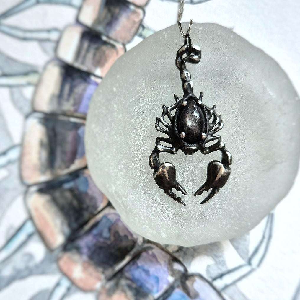 close-up of a silver Scorpion necklace with black gemstone, lying on a watercolor sketch