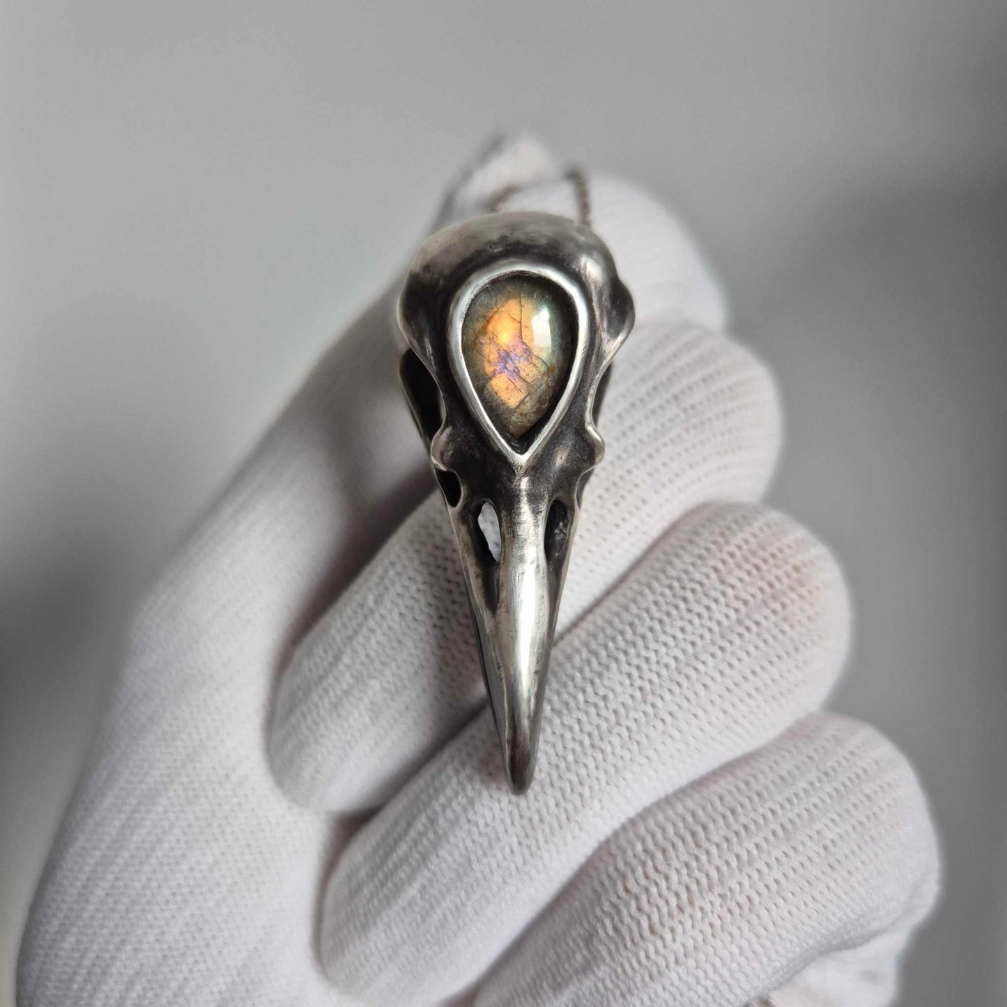 Silver raven skull necklace with golden-flash labradorite held in white glove, front view