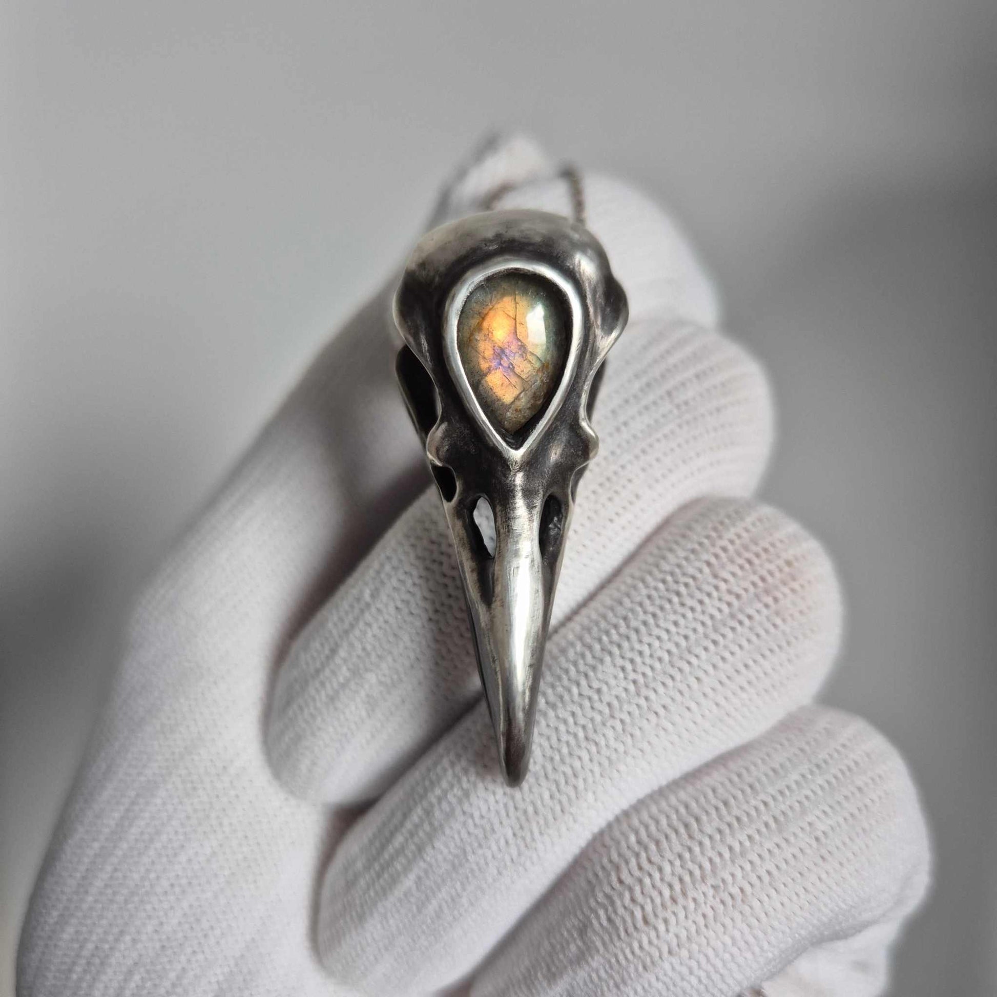 Silver raven skull necklace with golden-flash labradorite held in white glove, front view