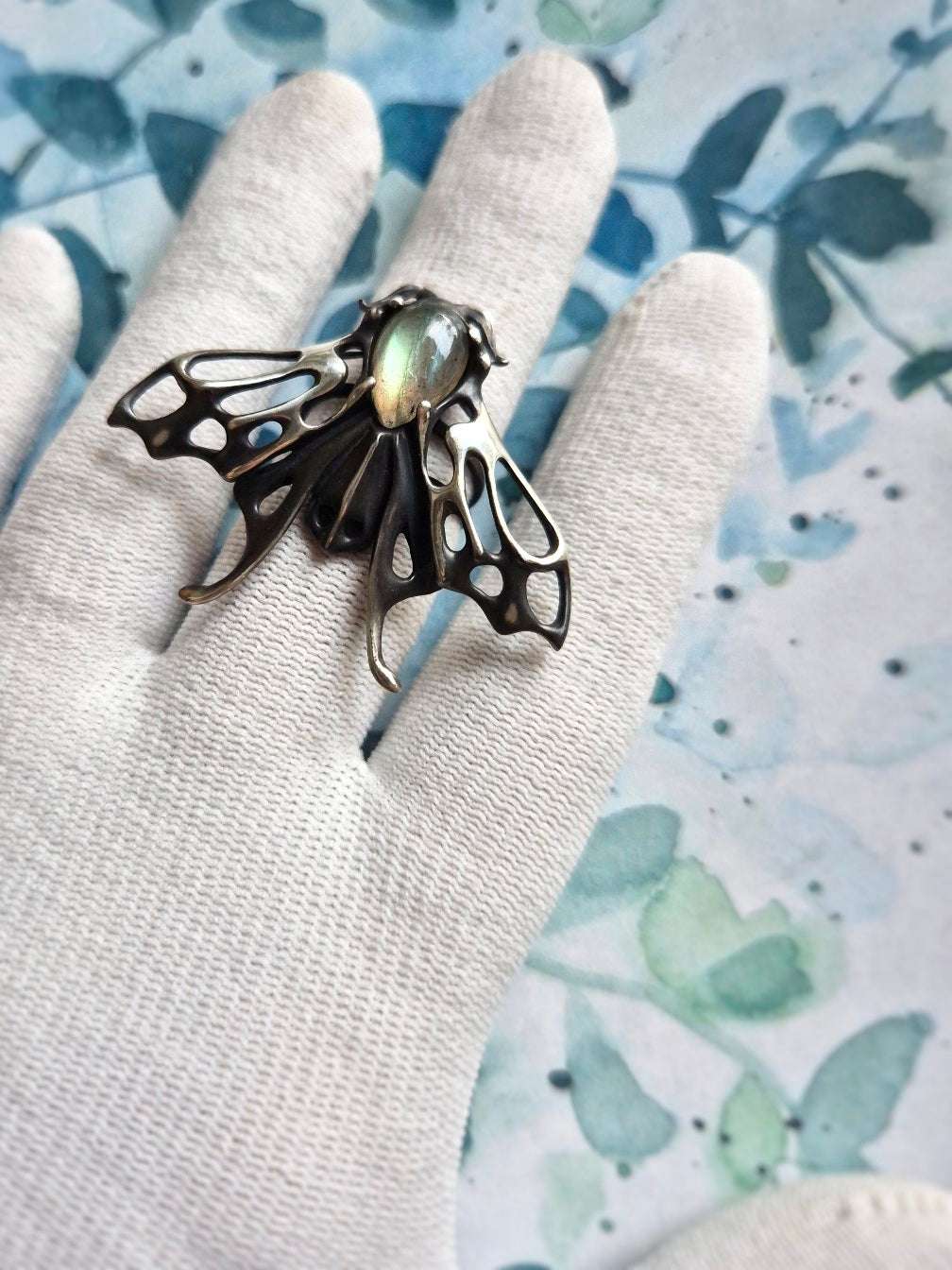 Hand holding silver Moth ring with shimmering labradorite gem, ethereal insect ring designHand holding silver Moth ring with shimmering labradorite gem, ethereal insect ring design, shown on botanical illustration background