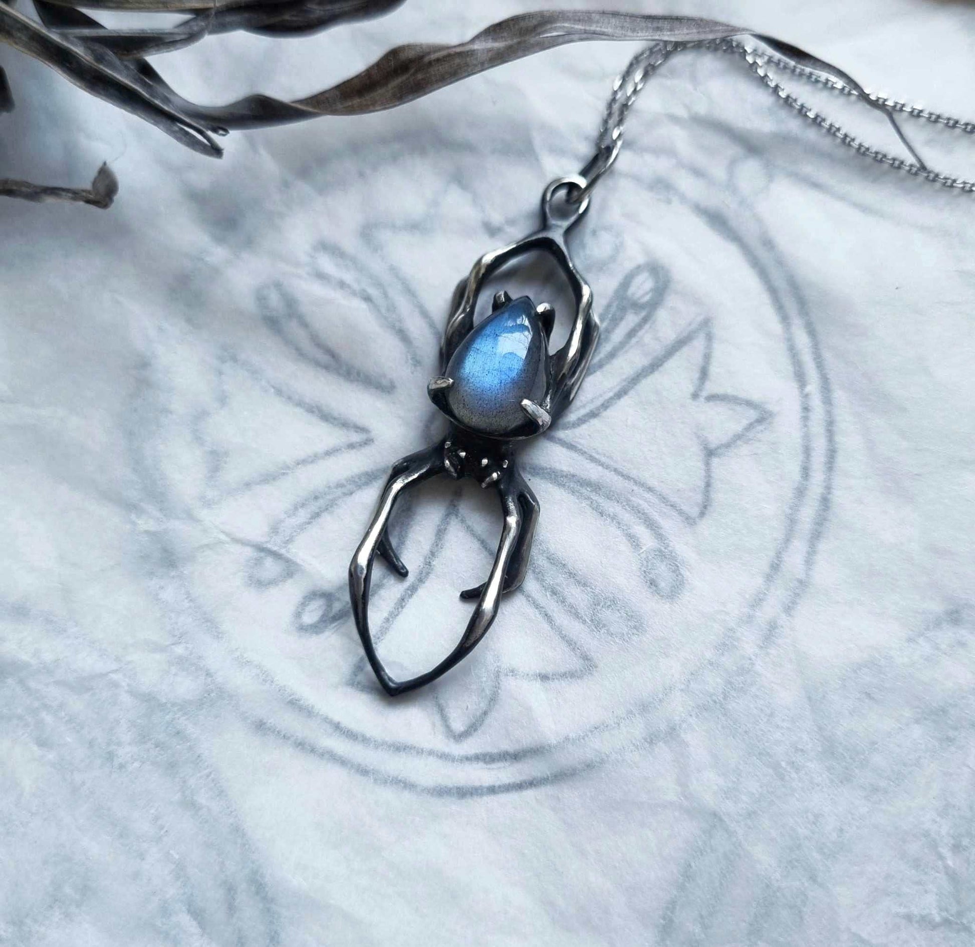 Silver Spider necklace with blue labradorite gemstone placed on a pattern pencil sketch, emphasizing symbolism and craftsmanship.