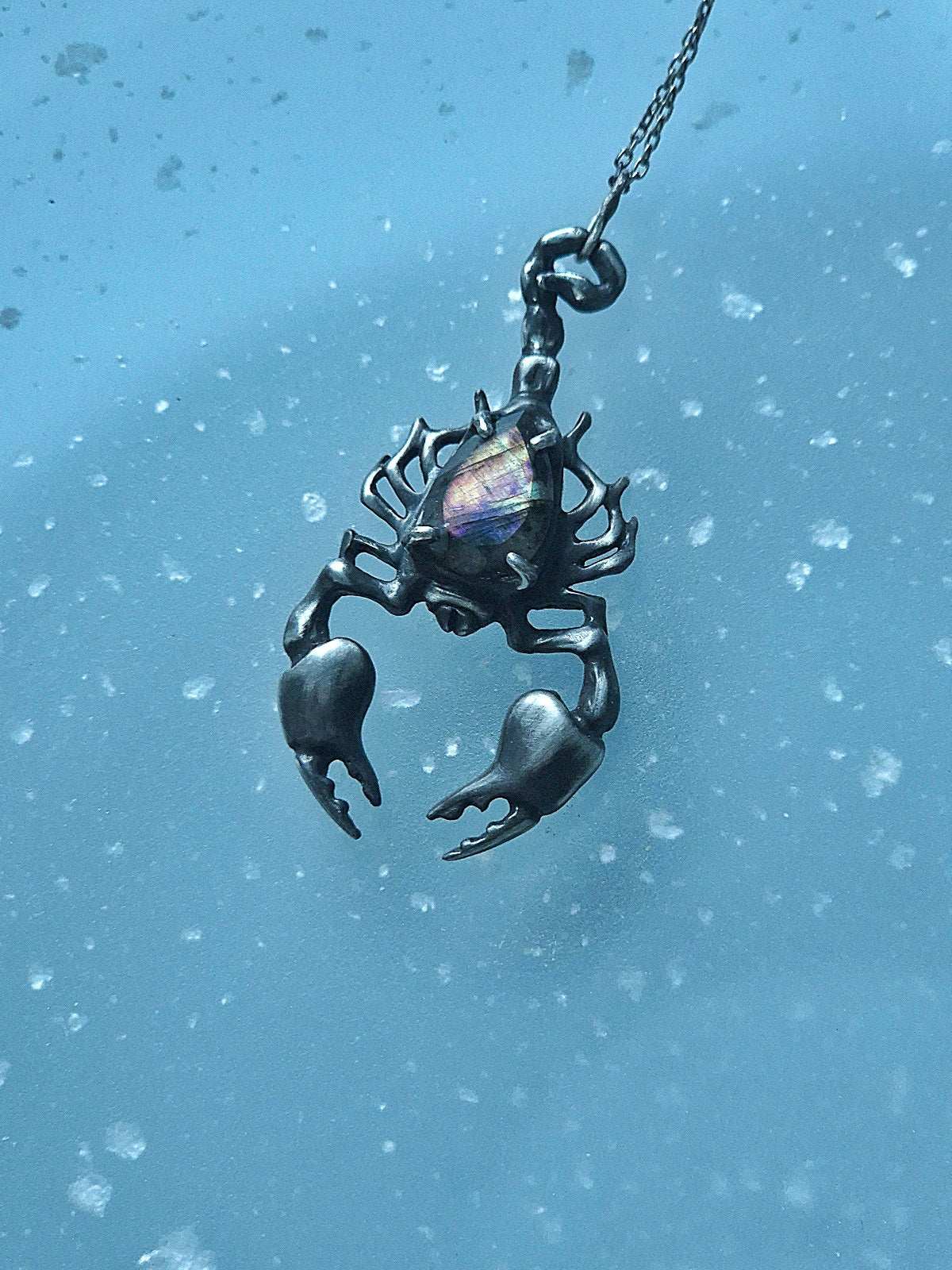 Silver Scorpion necklace with a faceted labradorite, resting on a softly speckled blue surface.