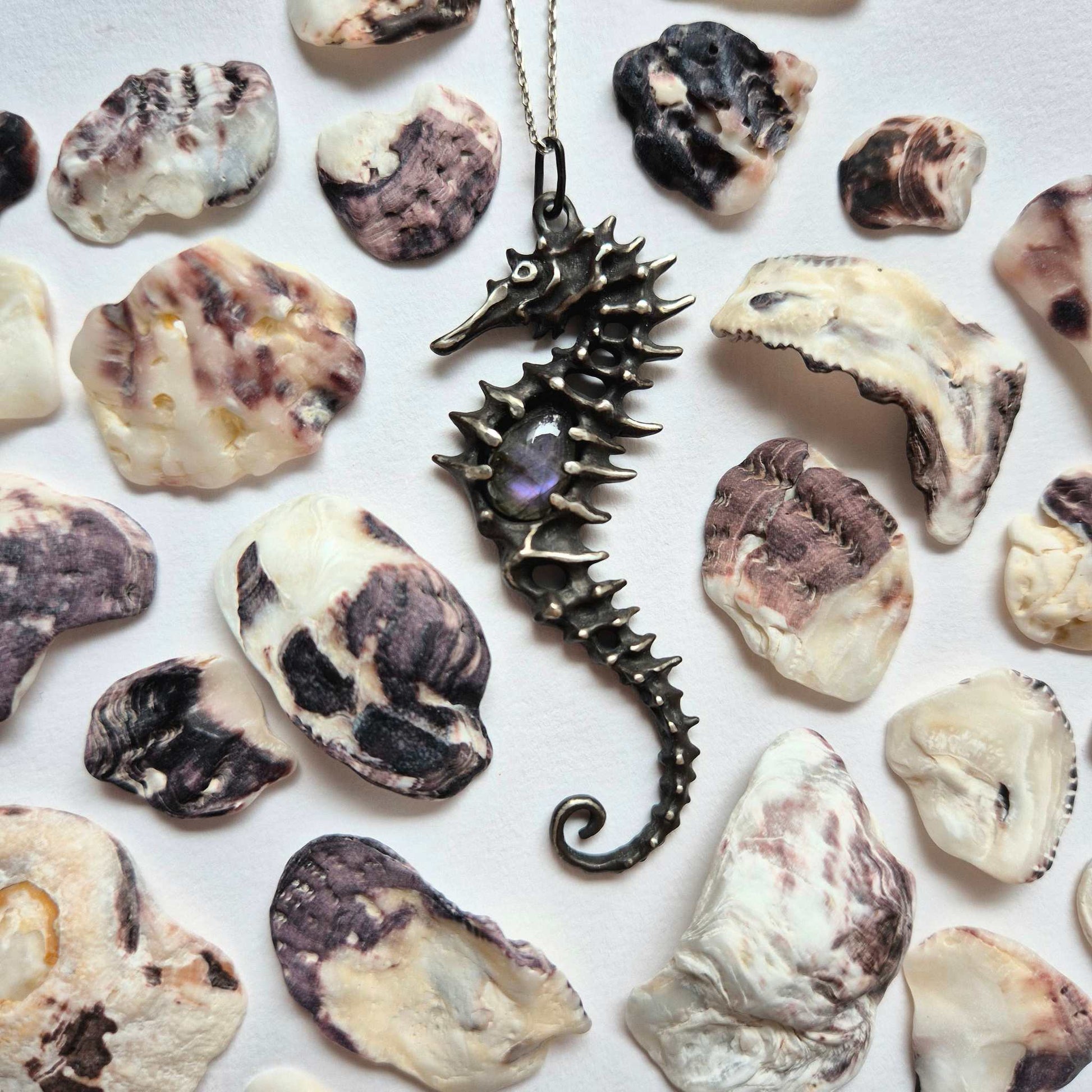 Silver seahorse necklace with purple labradorite placed among various natural seashells on a light surface