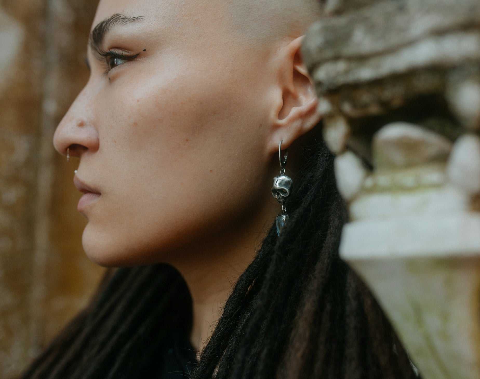 Close-up of handmade silver Skull earrings with labradorites, worn by a model with dreadlocks on textural background
