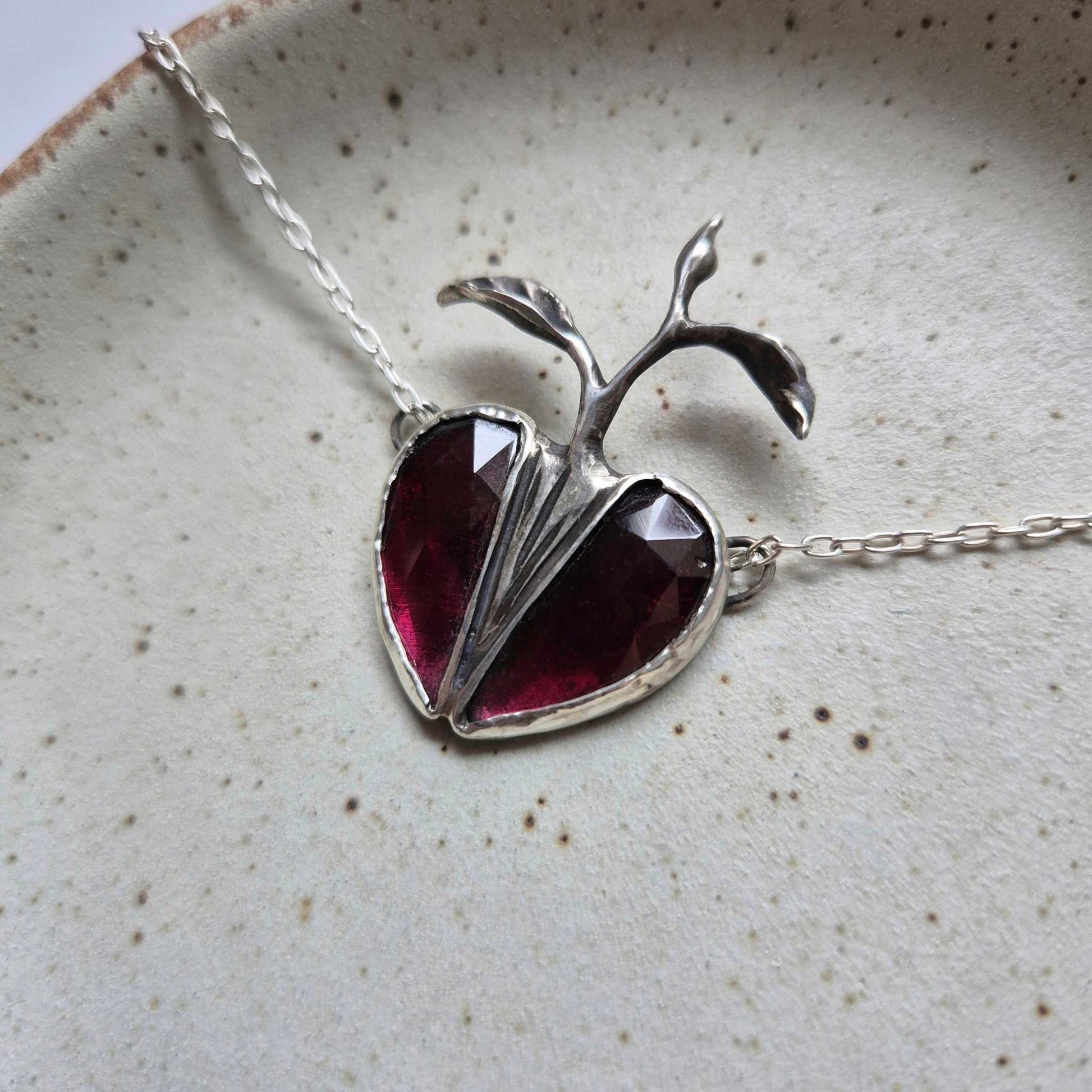 Silver Seed Heart necklace resting on soft white fabric, with the garnets glowing in ambient light.