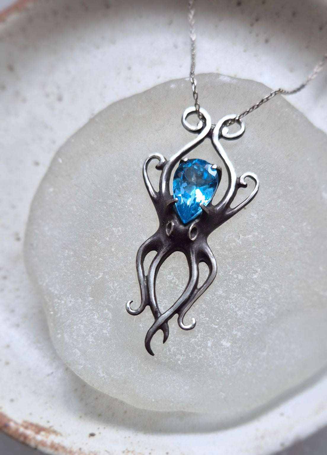 Close-up of silver Octopus necklace with faceted Swiss blue topaz, placed on sea glass