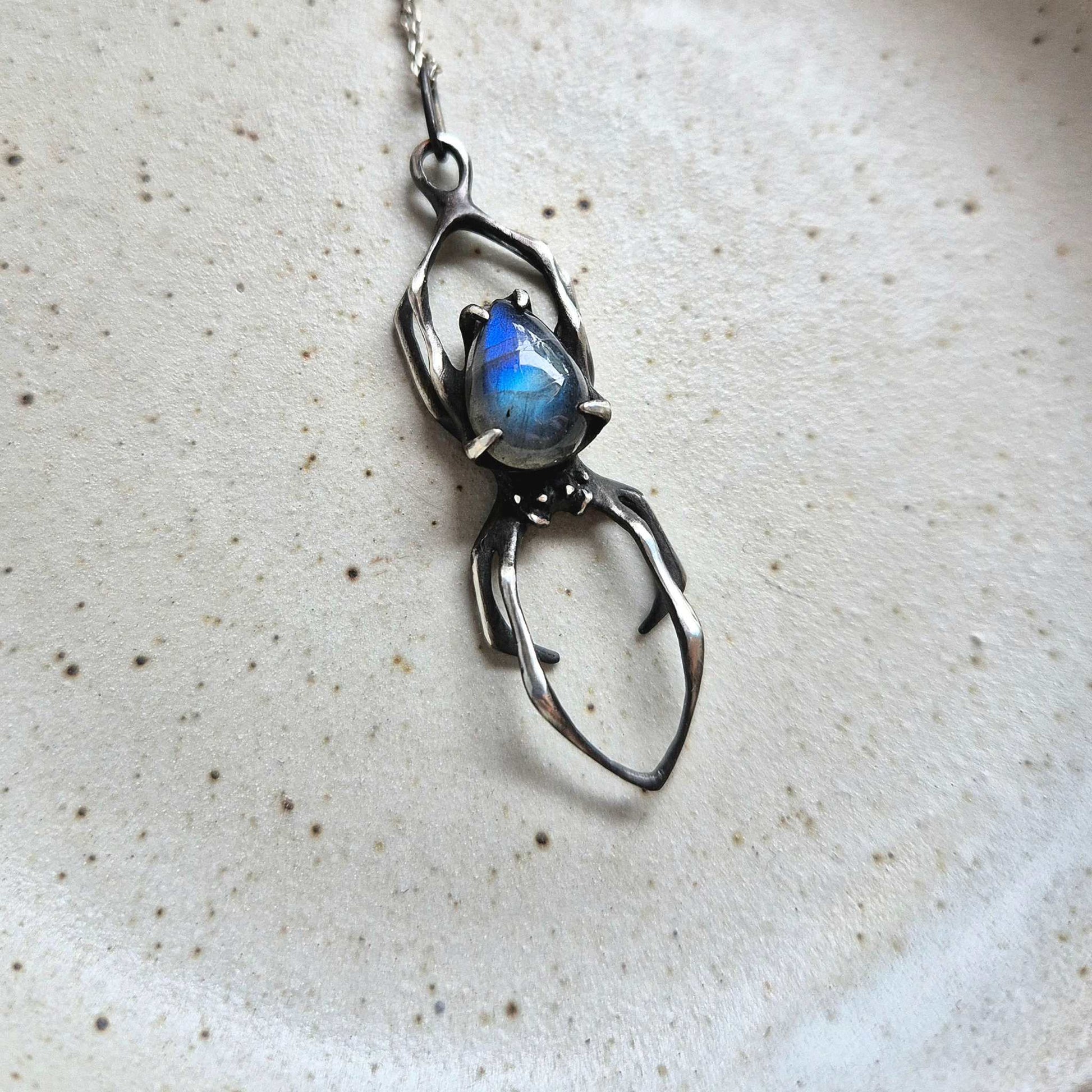 Close-up of silver Spider necklace featuring a glowing blue labradorite, displayed on a ceramic surface.