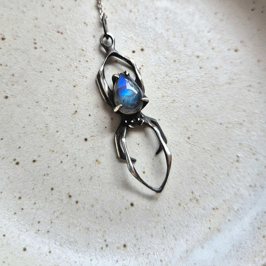 Close-up of silver Spider necklace featuring a glowing blue labradorite, displayed on a ceramic surface.
