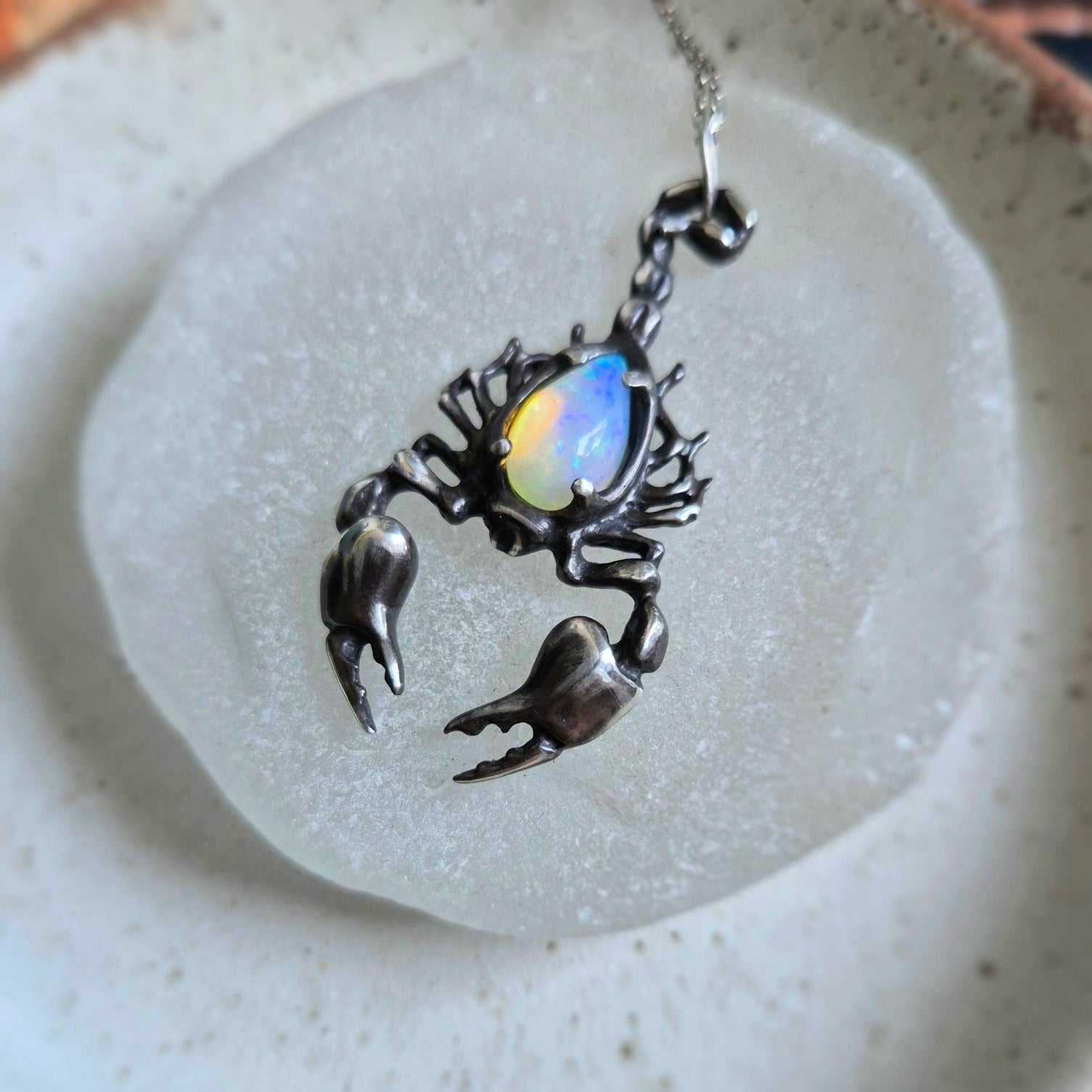 Angled shot of the silver Scorpion necklace with an opal gemstone, highlighting the contrast between the organic shape, oxidized silver and iridescent stone